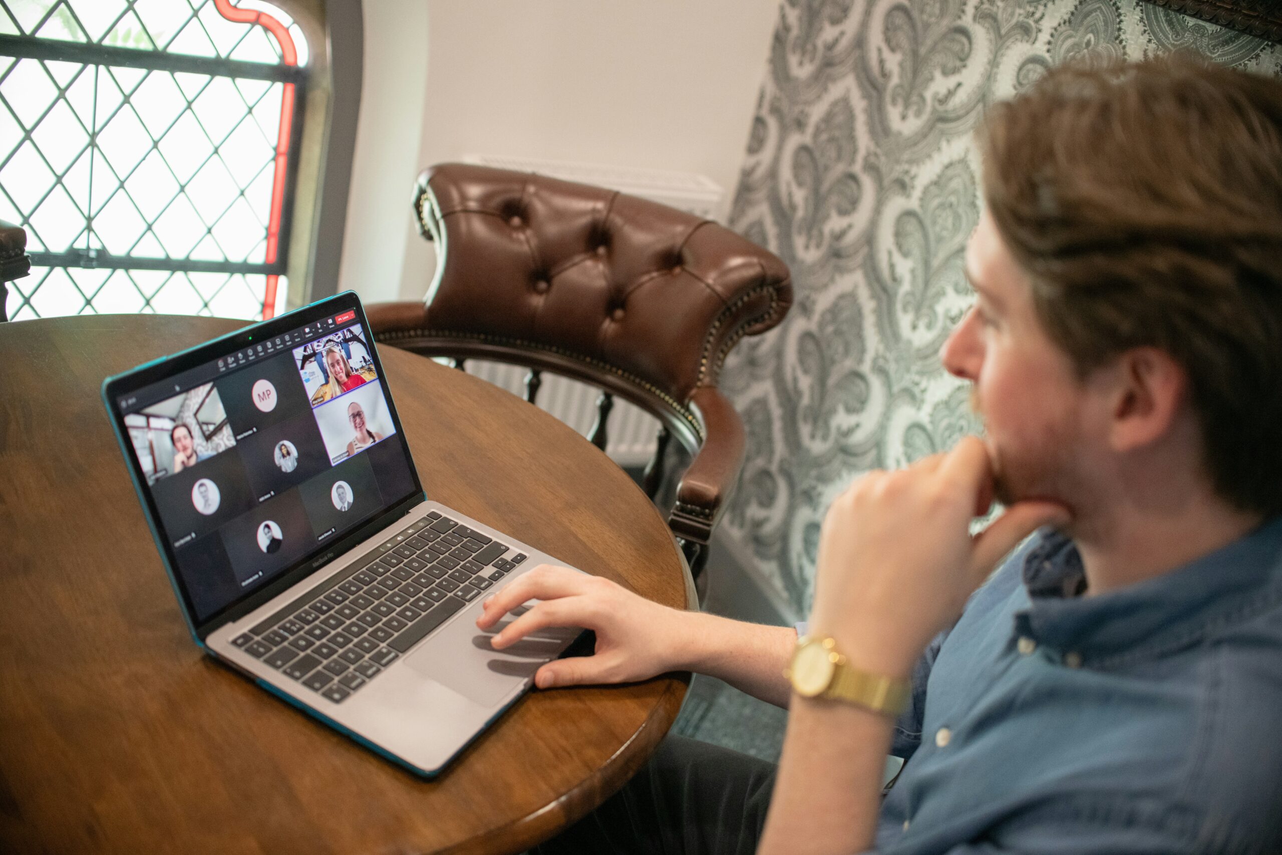 A laptop on a wooden table showing a group video call, with a participant's hand on the trackpad.
