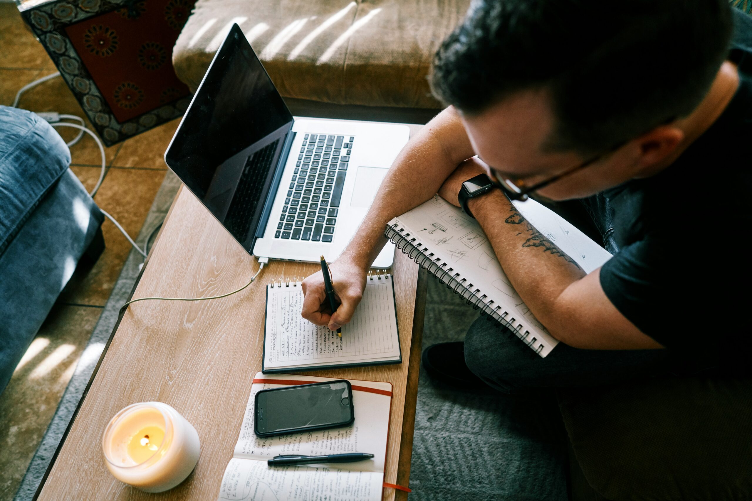 A young man writing in a notebook beside his open laptop, working through a plan at home.